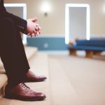 man in black pants and pair of brown leather lace-up shoes sitting on brown carpeted stairs inside room
