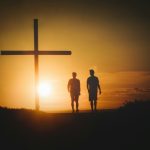 two people standing in front of a cross at sunset